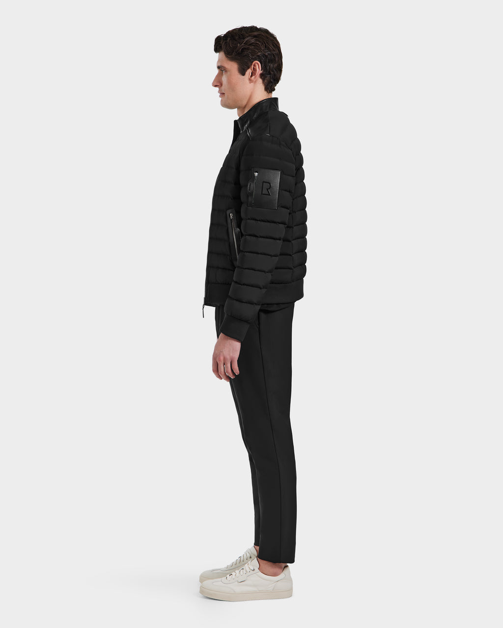 A man stands in profile against a plain background, wearing the Rudsak JOSHUA - 6124542 Black mens biker down jacket, paired with black pants and white sneakers.