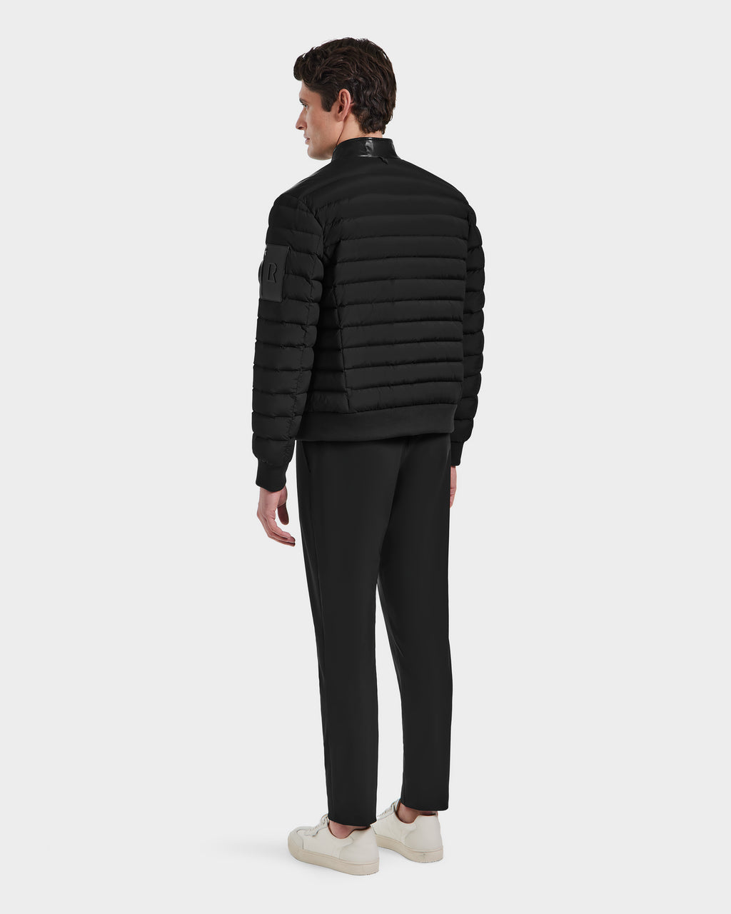 A man wearing the Rudsak JOSHUA - 6124542 Black quilted light down jacket with a leather collar, black pants, and white sneakers stands facing away against a plain light background.