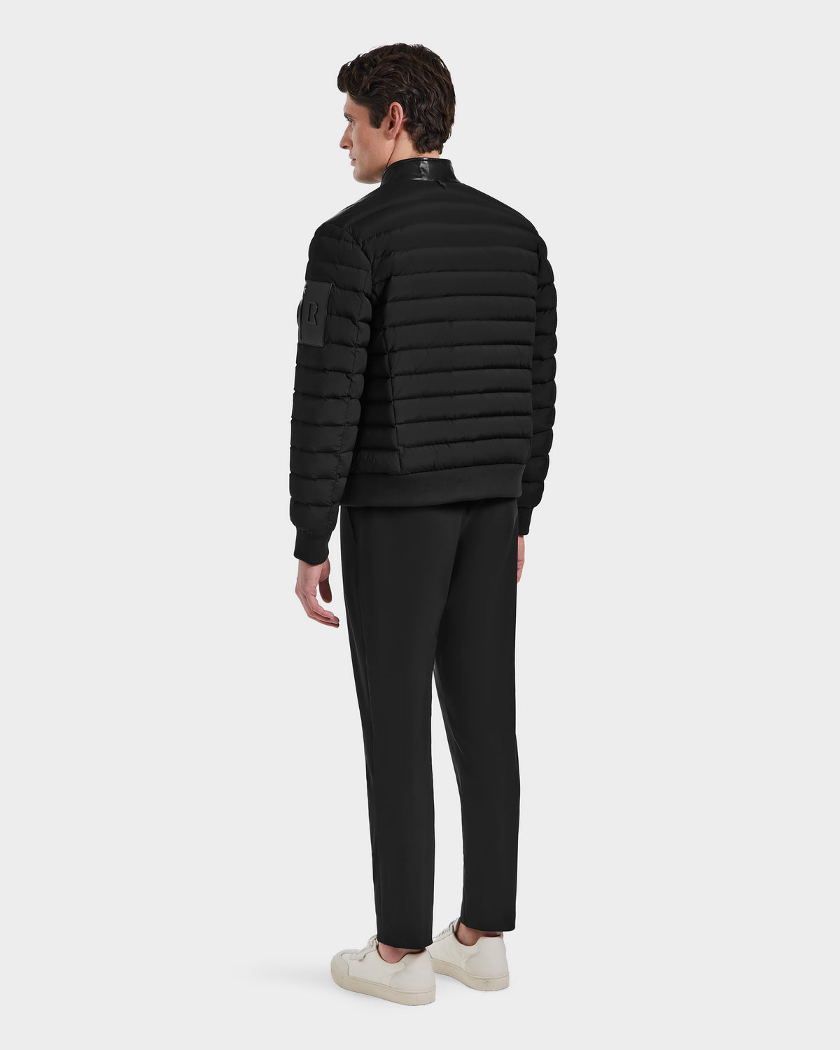 A man wearing the Rudsak JOSHUA - 6124542 Black quilted light down jacket with a leather collar, black pants, and white sneakers stands facing away against a plain light background.