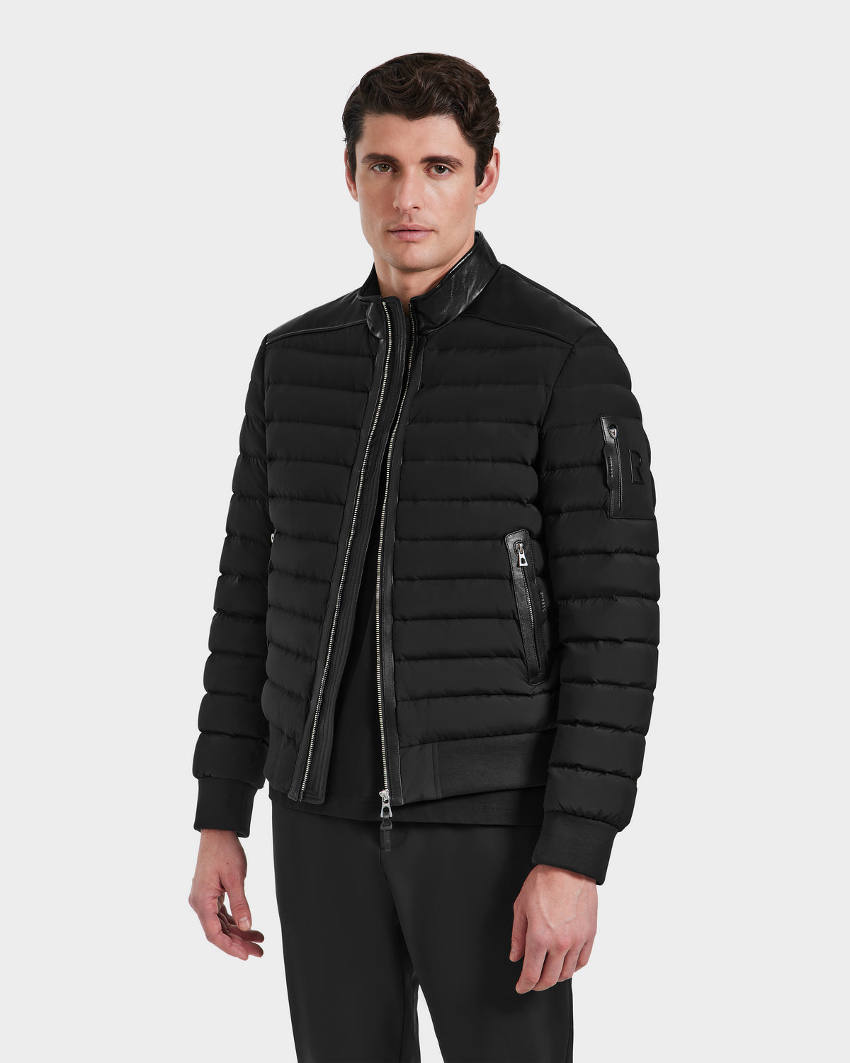 A man stands facing forward against a plain white background, wearing Rudsaks JOSHUA - 6124542 Black quilted light down jacket with a zipper and standing collar, paired with black pants.