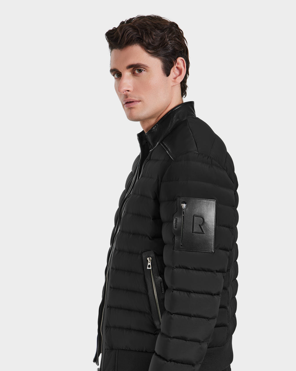 A young man with short brown hair models the Rudsak JOSHUA - 6124542 Black mens biker down jacket, featuring a zip pocket and leather patch on the sleeve, while looking sideways against a plain light background.