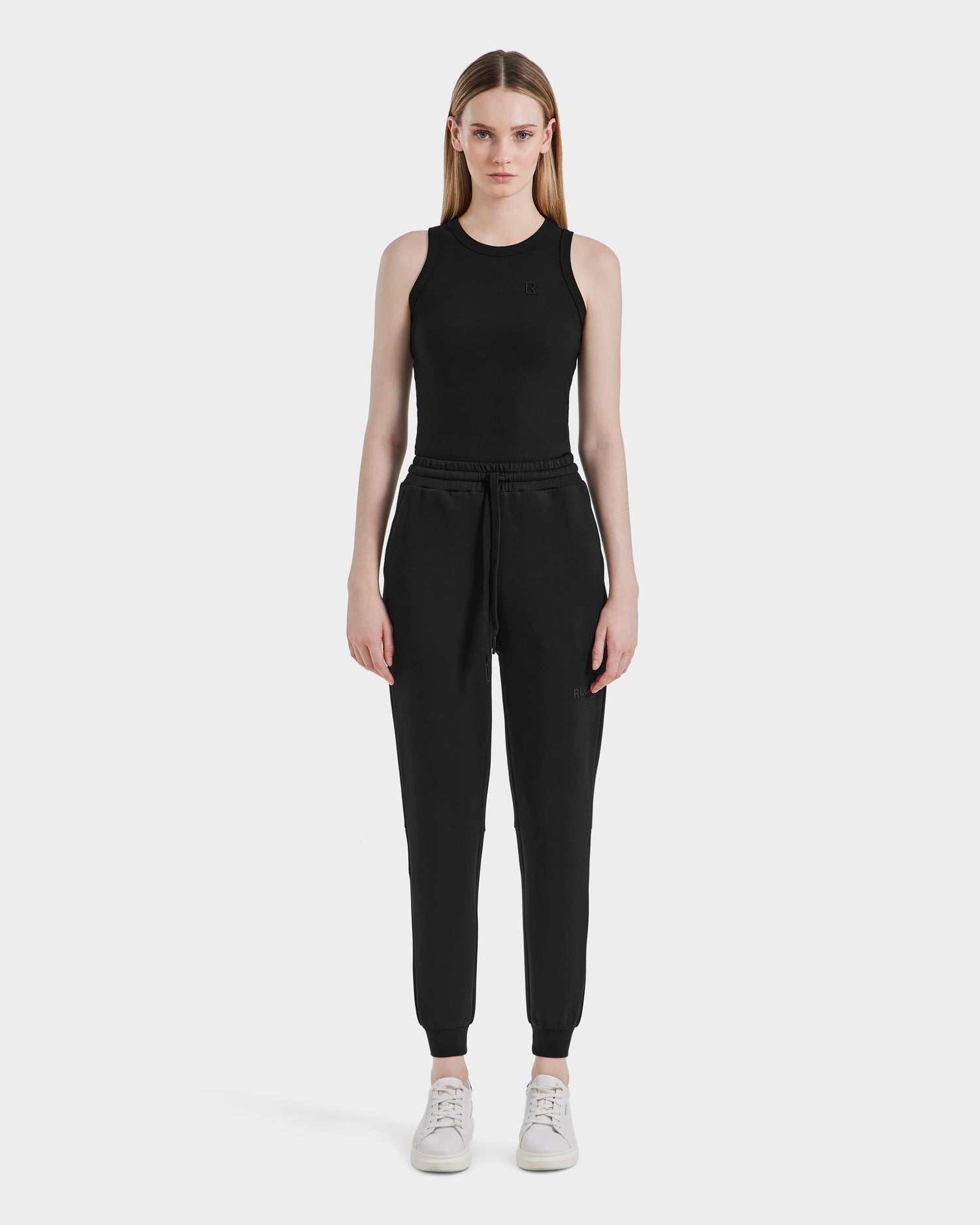 A woman faces forward against a white background, wearing Rudsaks JOSS - 9524009 Black W sleeveless top and stylish cotton joggers with a drawstring waist, paired with white sneakers.
