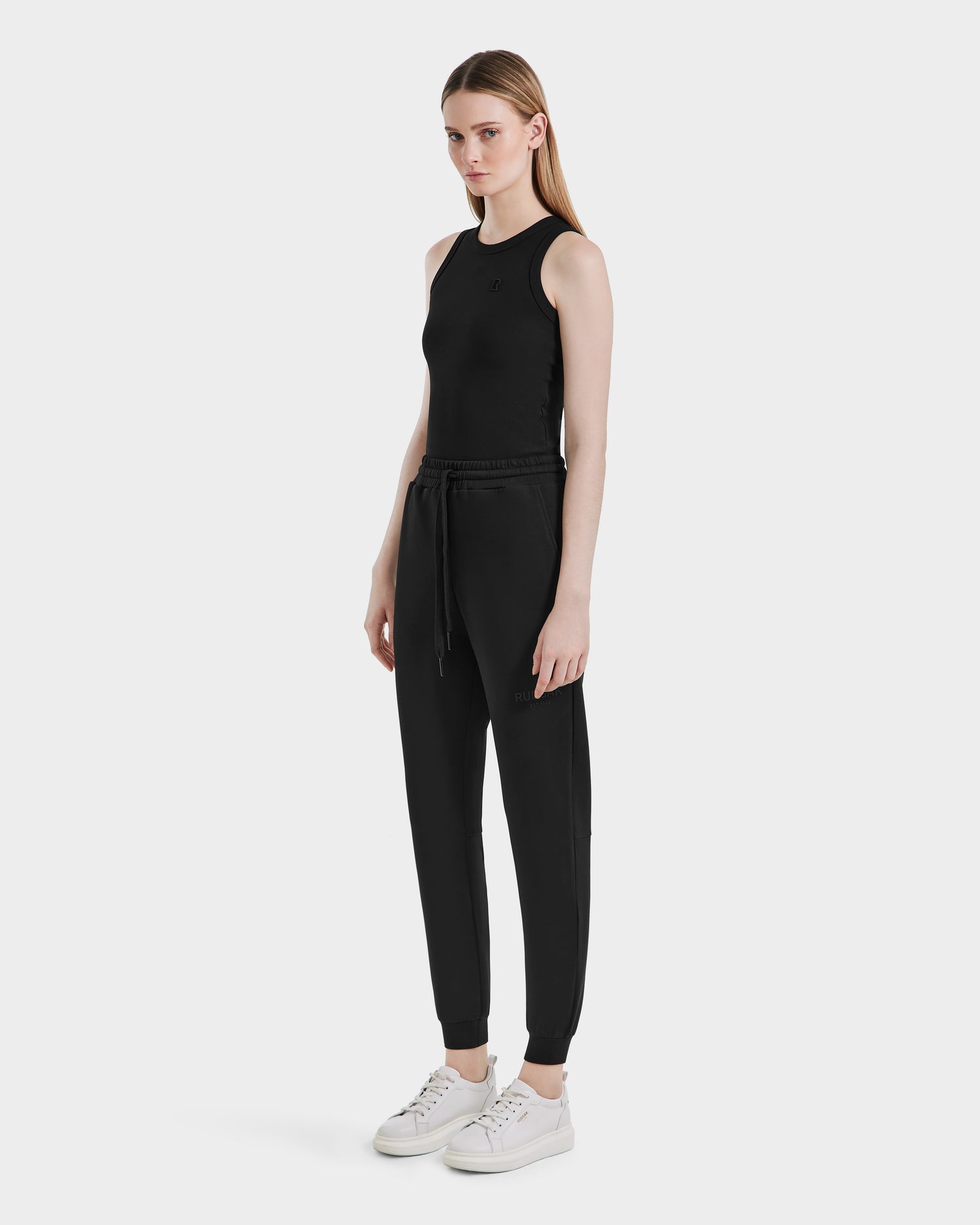 A woman faces forward, wearing the Rudsak JOSS - 9524009 Black W sleeveless top and stylish joggers with a drawstring waist, paired with white sneakers. She has straight light brown hair and a neutral expression against a plain white background.