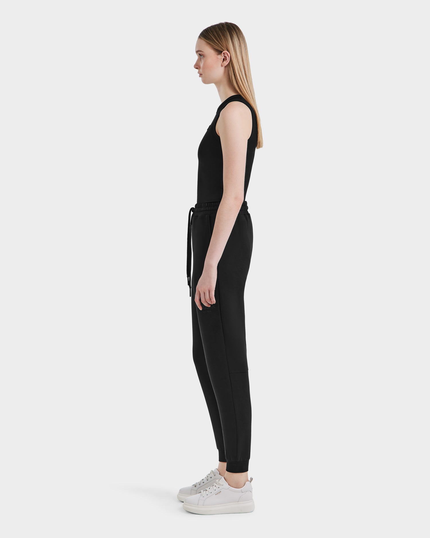 A young woman with straight light brown hair and a neutral expression stands in profile, wearing the Rudsak JOSS - 9524009 Black W sleeveless jumpsuit with a drawstring waist and white sneakers against a plain background.