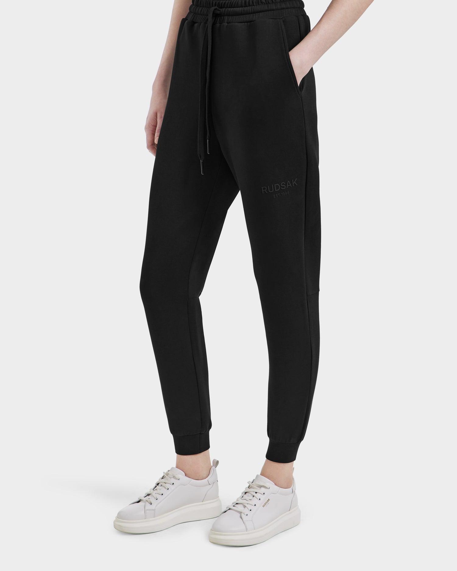 A person wears Rudsaks JOSS - 9524009 Black W premium cotton joggers with RUDSAK branding on the left thigh, paired with white sneakers and a white top. One hand is in their pocket, standing out against a plain white background.