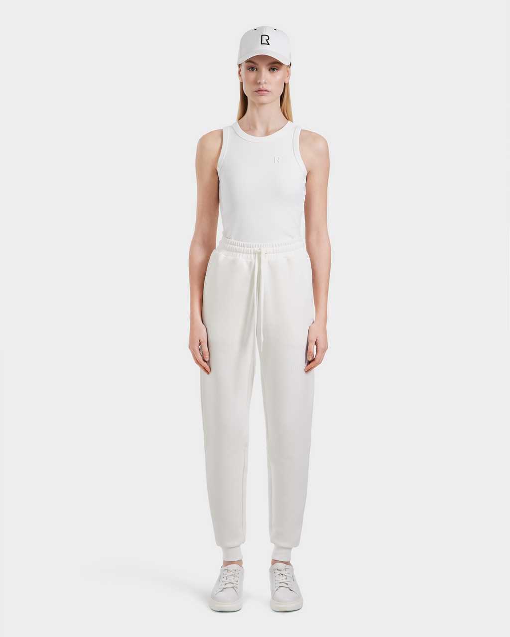 A person stands facing forward against a white background, wearing Rudsak JOSS - 9524009 Iceland W premium cotton blend unisex jogger pants, a white tank top, white sneakers, and a white baseball cap.