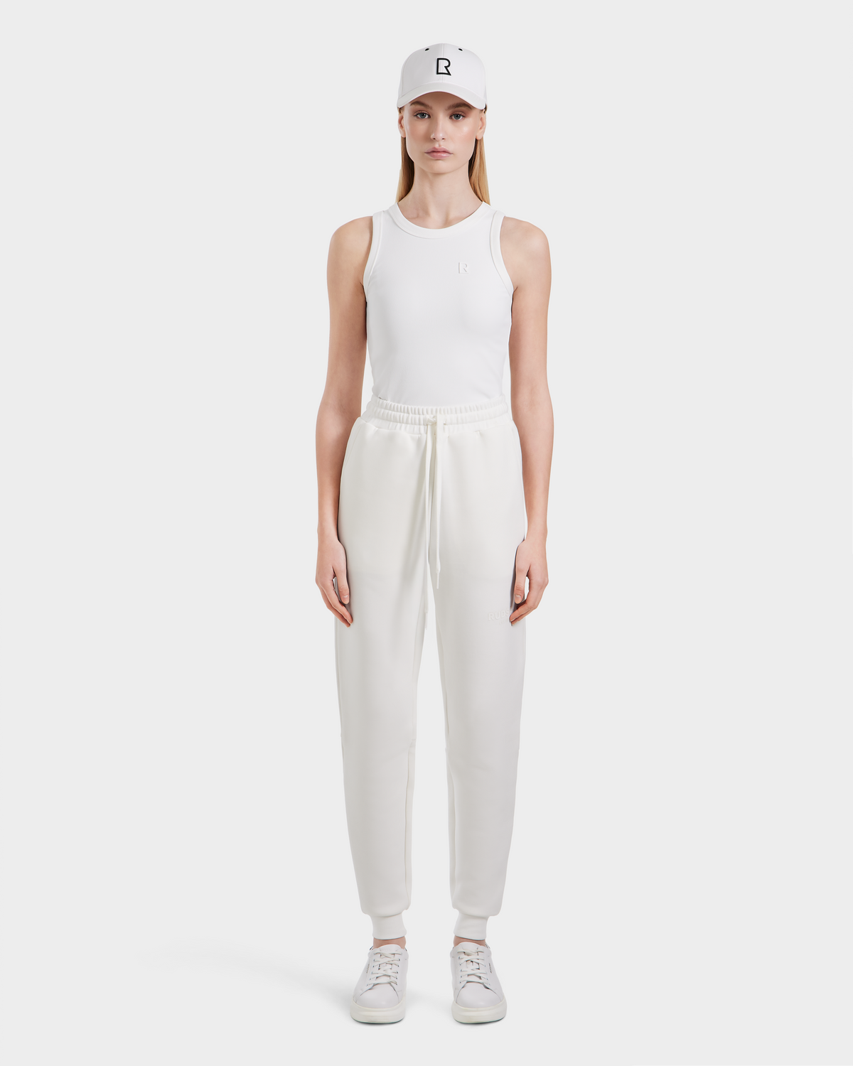A person stands facing forward against a white background, wearing Rudsak JOSS - 9524009 Iceland W premium cotton blend unisex jogger pants, a white tank top, white sneakers, and a white baseball cap.