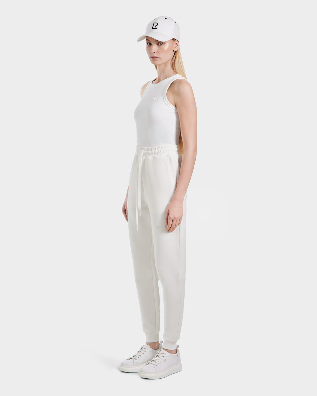 A woman with long blonde hair stands sideways against a plain background, wearing Rudsaks JOSS - 9524009 Iceland W: a white cap, sleeveless top, modern premium cotton jogger pants, and white sneakers.