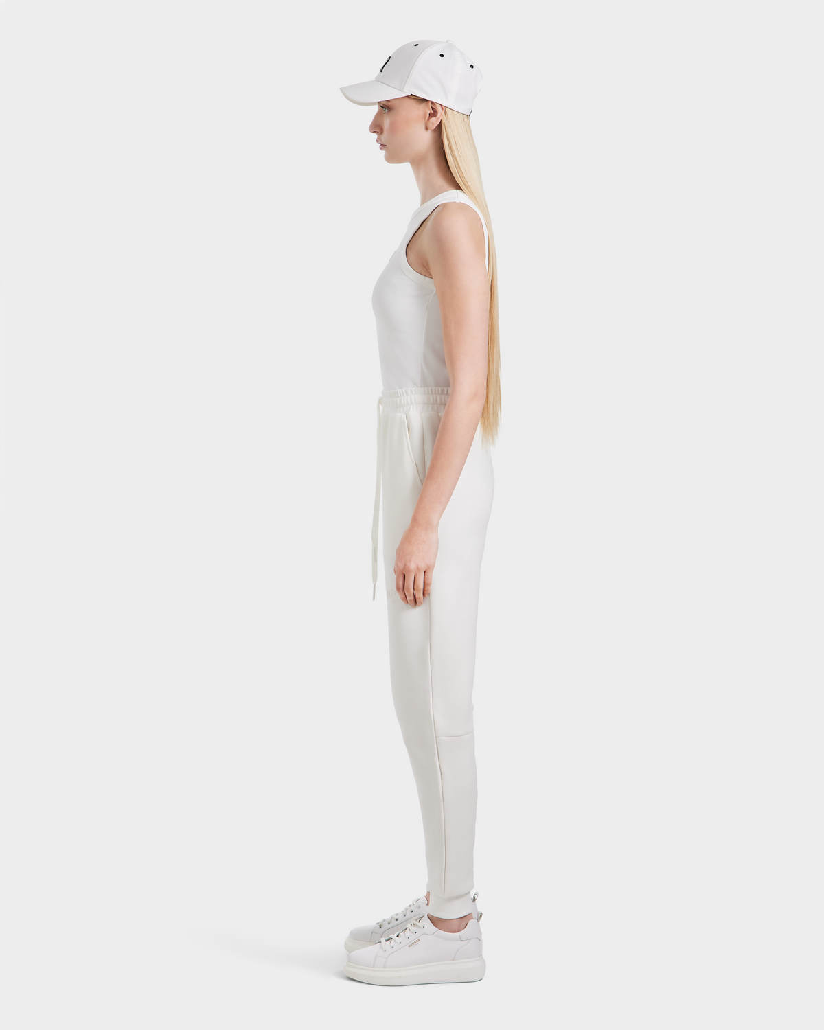 A woman with long blonde hair stands in profile against a white background, wearing the Rudsak JOSS - 9524009 Iceland W premium cotton blend white tank top, modern joggers, white sneakers, and a matching baseball cap.