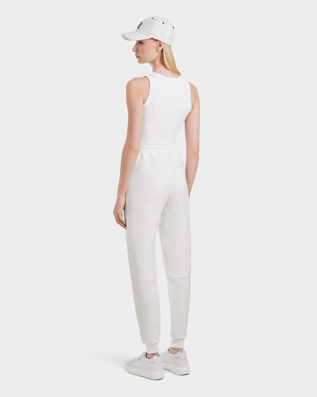 A person stands facing away, wearing Rudsak JOSS - 9524009 Iceland W jogger pants with a white sleeveless top, white sneakers, and a white cap against a plain light background.
