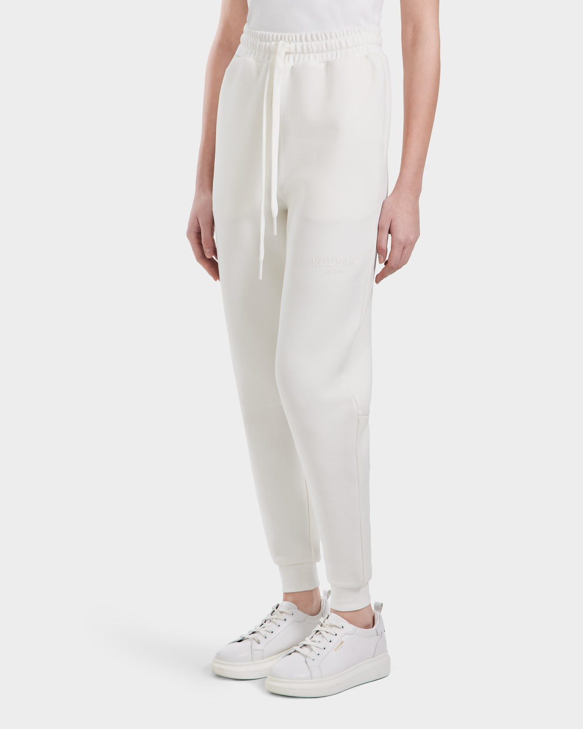 A person showcases the Rudsak JOSS - 9524009 Iceland W premium cotton joggers with a drawstring waist and white sneakers, standing against a plain white background. The image focuses on the modern joggers’ lower-half style.