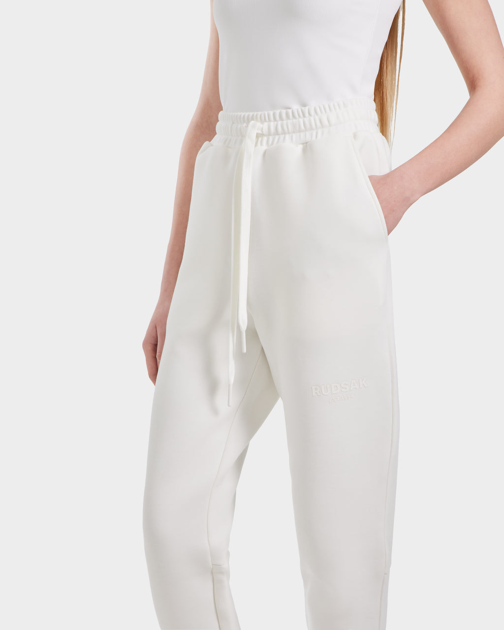Someone wears Rudsak’s JOSS - 9524009 Iceland W high-waisted white jogger sweatpants, crafted in a premium cotton blend with an elastic drawstring waistband, subtle RUDSAK branding on the left thigh, and a hand in the pocket.
