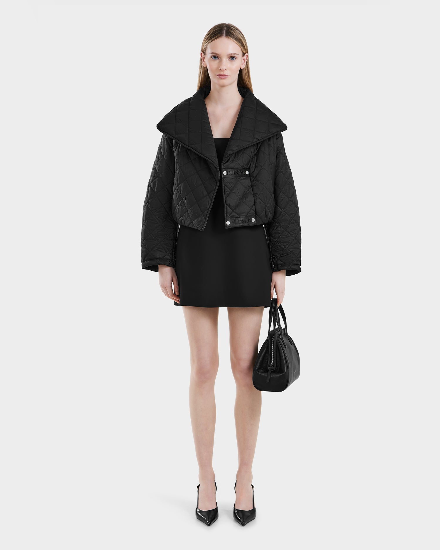A woman stands against a plain white background wearing the Rudsak KALEE - 8124005 Black cropped quilted jacket, paired with a short black dress, black pointed heels, and carrying a black handbag.
