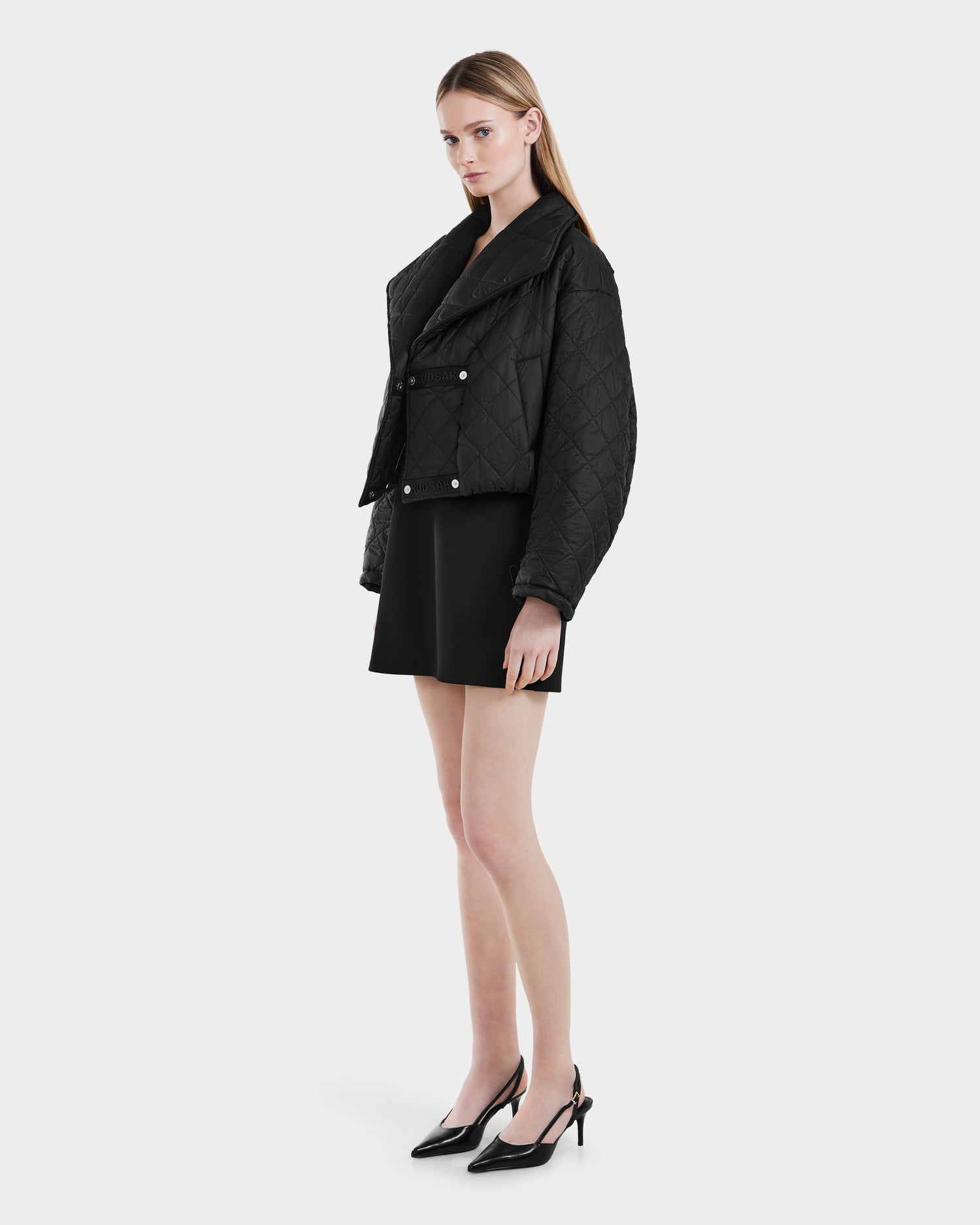 A woman stands against a plain light background wearing the Rudsak KALEE - 8124005 Black womens cropped quilted jacket, styled with a black mini skirt and pointed-toe heels. She gazes slightly to the side with a neutral expression.