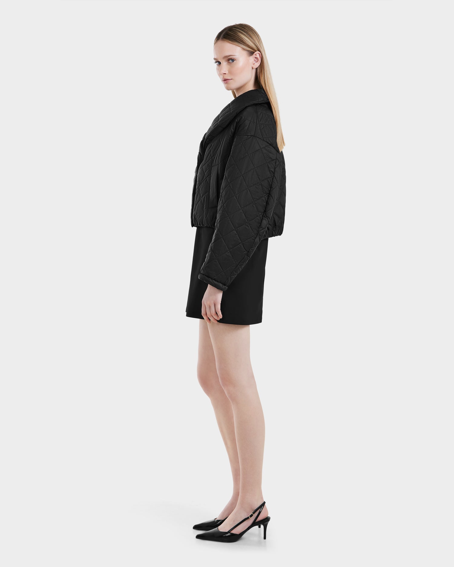 A woman with straight blonde hair stands sideways, wearing the Rudsak KALEE - 8124005 Black womens cropped quilted light jacket with a black skirt and slingback heels against a plain white background.