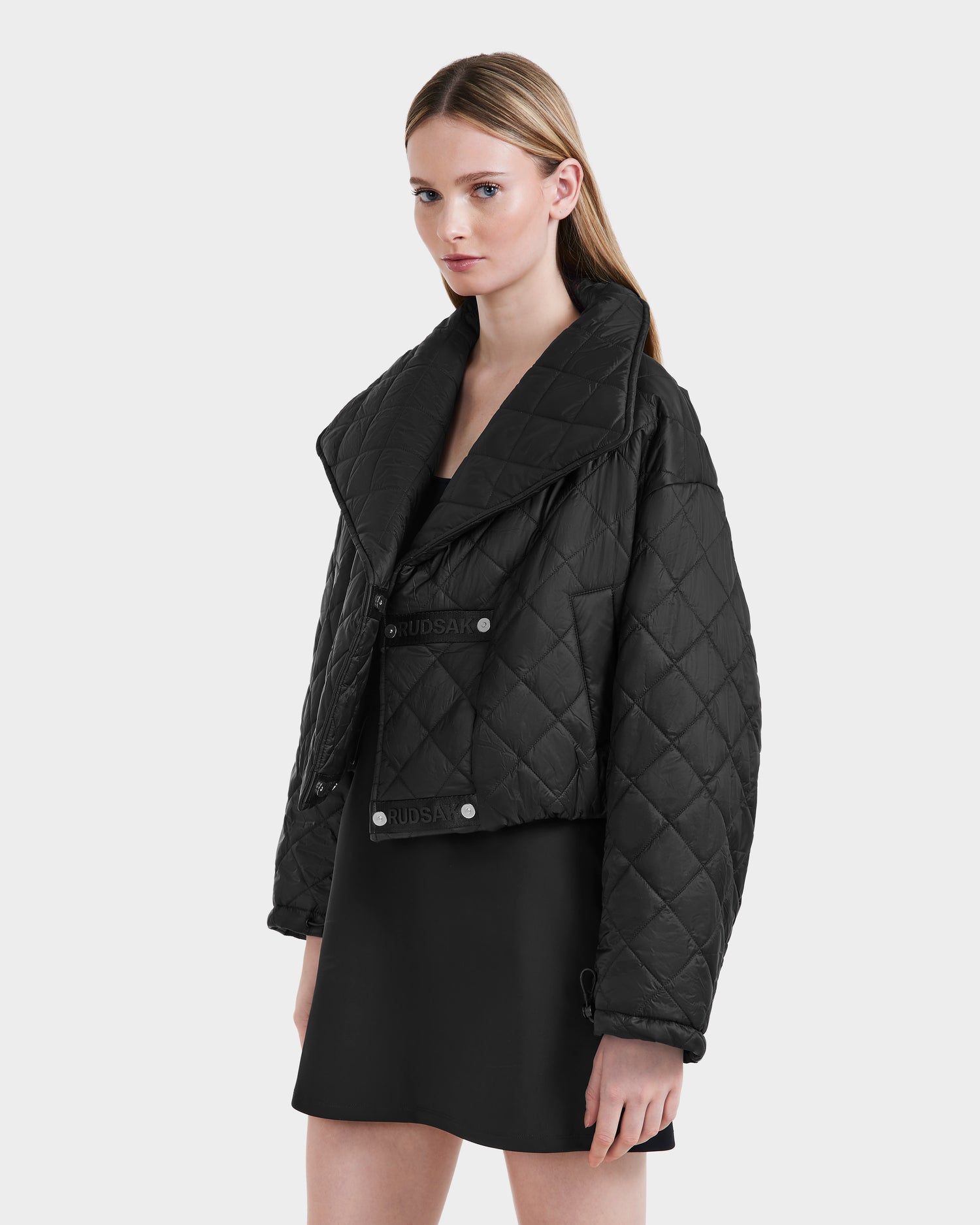 A woman with straight, light brown hair wears the Rudsak KALEE - 8124005 Black oversized quilted jacket with wide lapels over a short black dress, standing against a plain white background.