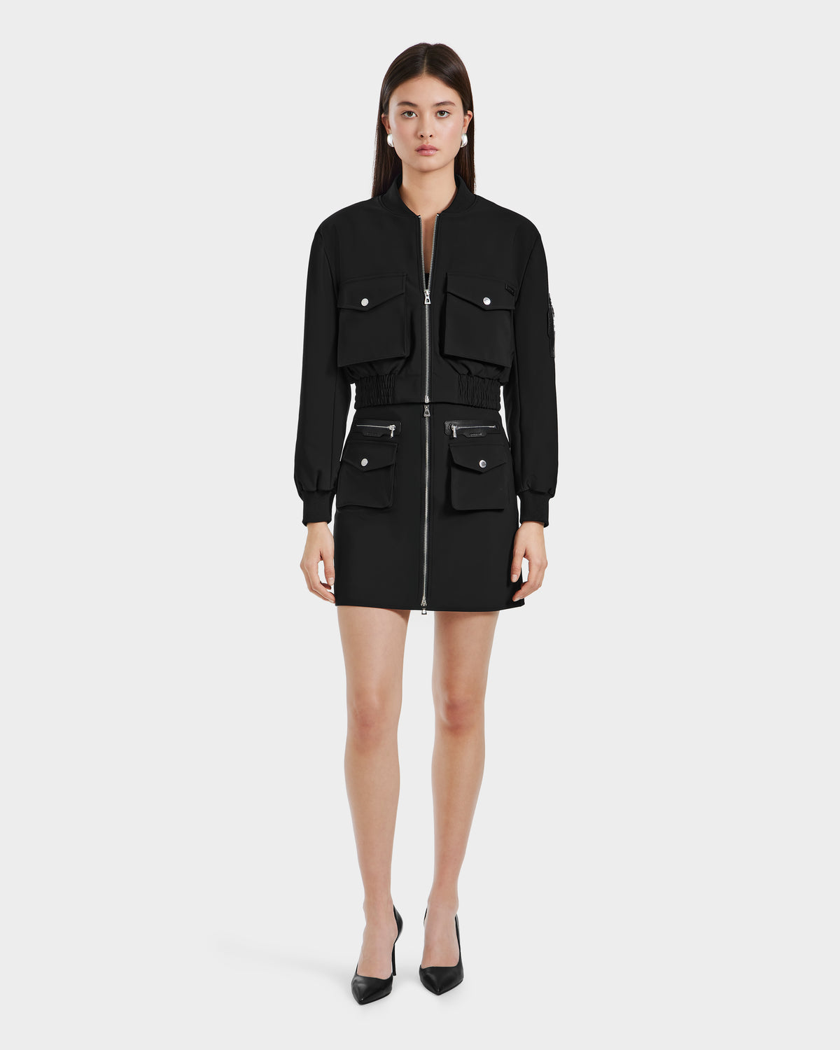 A woman faces forward against a white background, wearing the KEIRA - 8525024 Black dress by Rudsak with long sleeves, zip-up front, and multiple pockets, layered under a cropped bomber jacket and paired with black pointed heels.