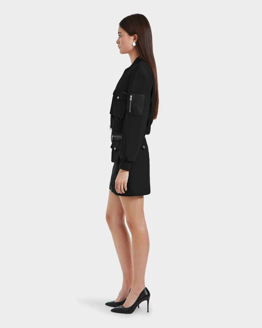 A woman with long brown hair stands in profile against a plain light background, wearing the Rudsak KEIRA - 8525024 Black lightweight jacket with pockets, paired with a black skirt and black high heels.