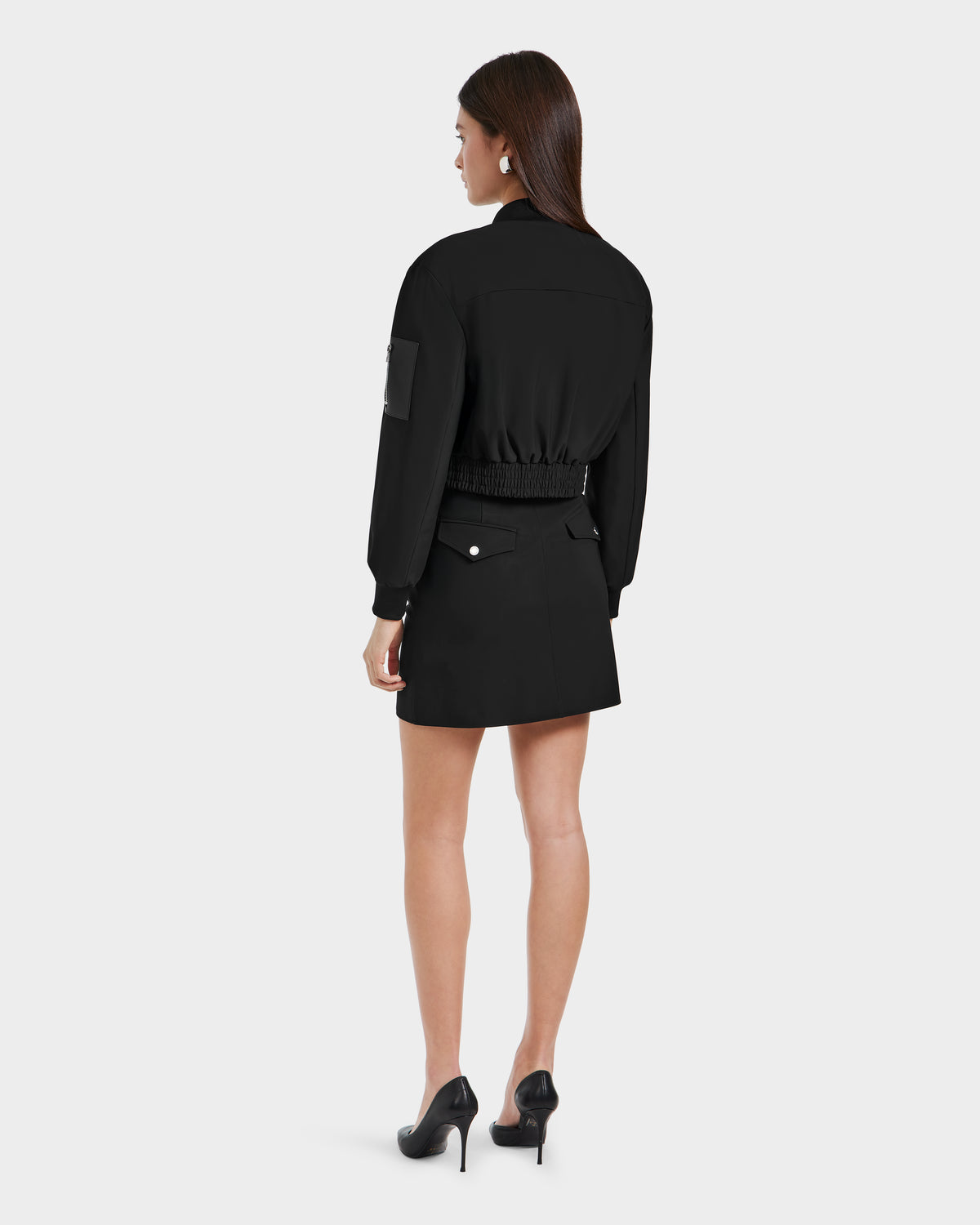 A woman with long brown hair faces away, wearing Rudsaks KEIRA - 8525024 Black vegan leather jacket with an elastic waistband and a matching skirt, paired with black heels against a plain light background.