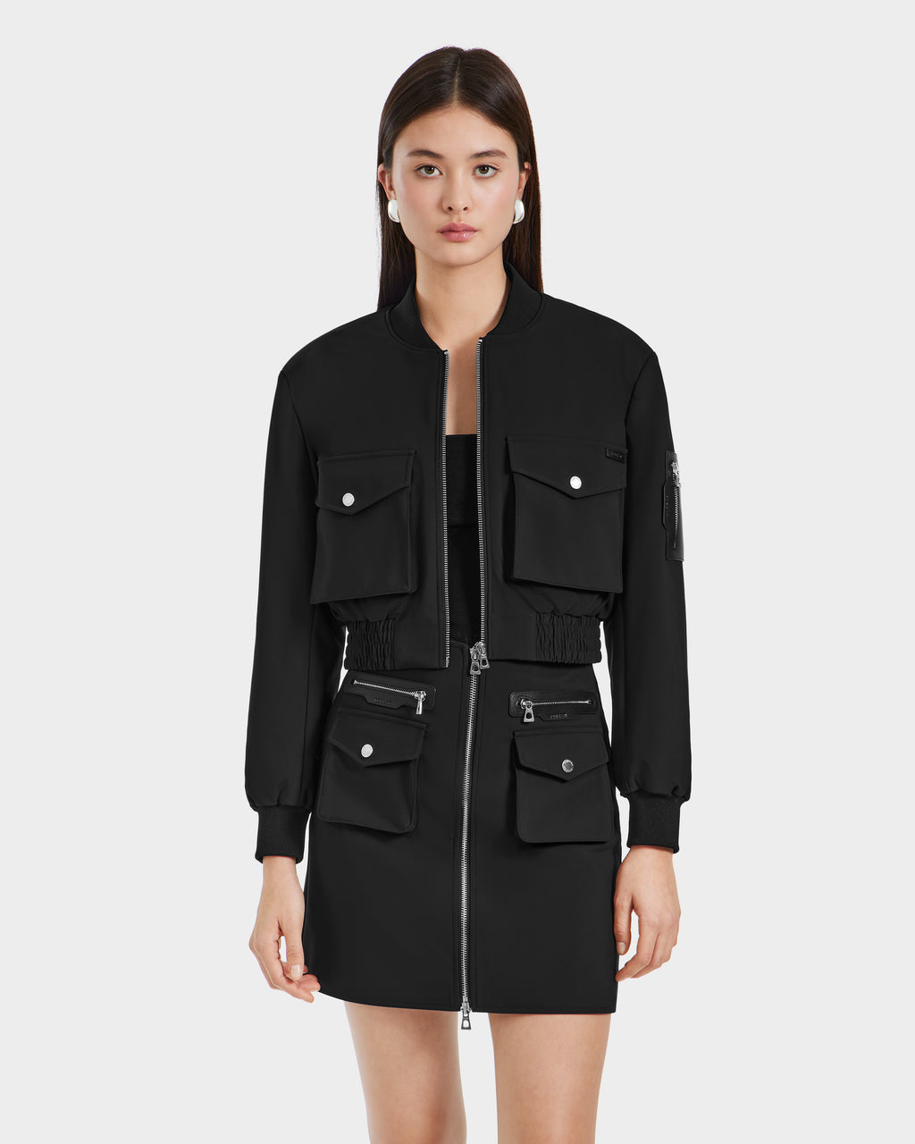 A woman with long dark hair wears the Rudsak KEIRA - 8525024 Black cropped bomber jacket and matching skirt, both with multiple front pockets and silver zippers, standing against a plain white background.
