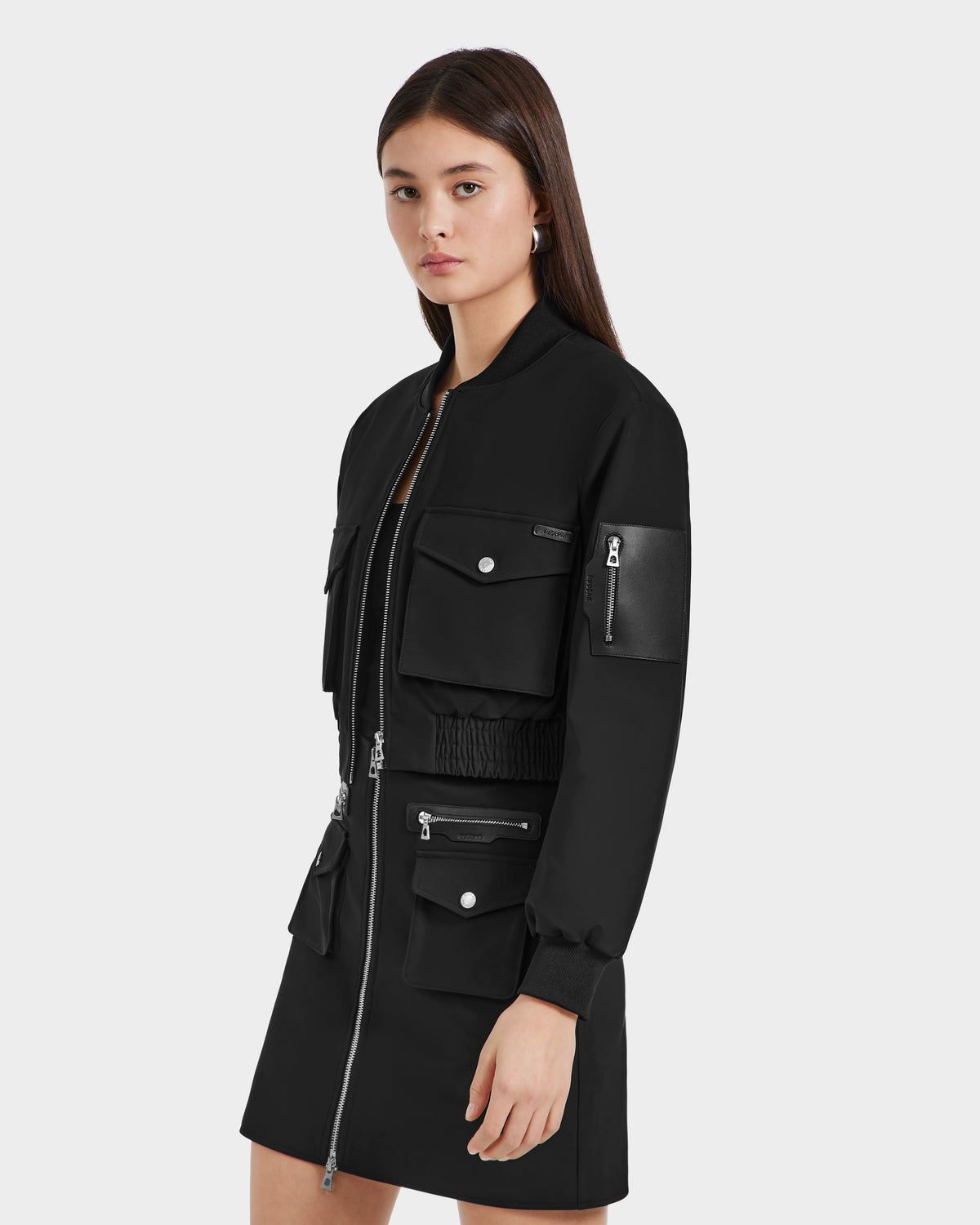 A woman with straight brown hair models the Rudsak KEIRA - 8525024 Black vegan leather utility jacket and matching skirt, featuring multiple pockets and silver zippers, against a plain light background.