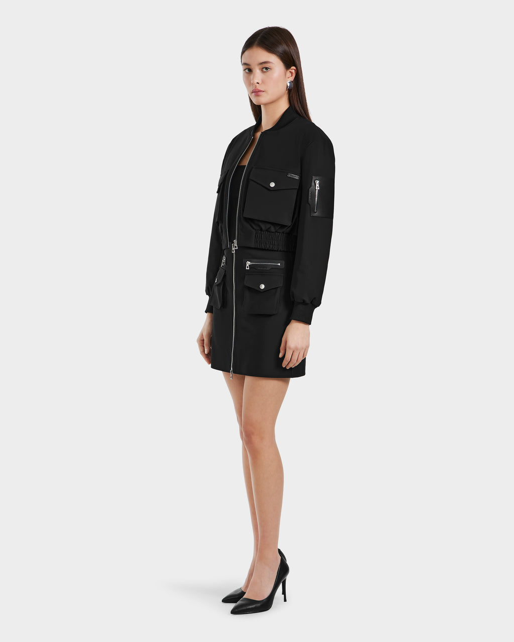 A woman stands in profile against a plain background, wearing the KEIRA - 8525024 Black zip-up dress by Rudsak with multiple front pockets and long sleeves, styled with a cropped bomber jacket and black high heels.