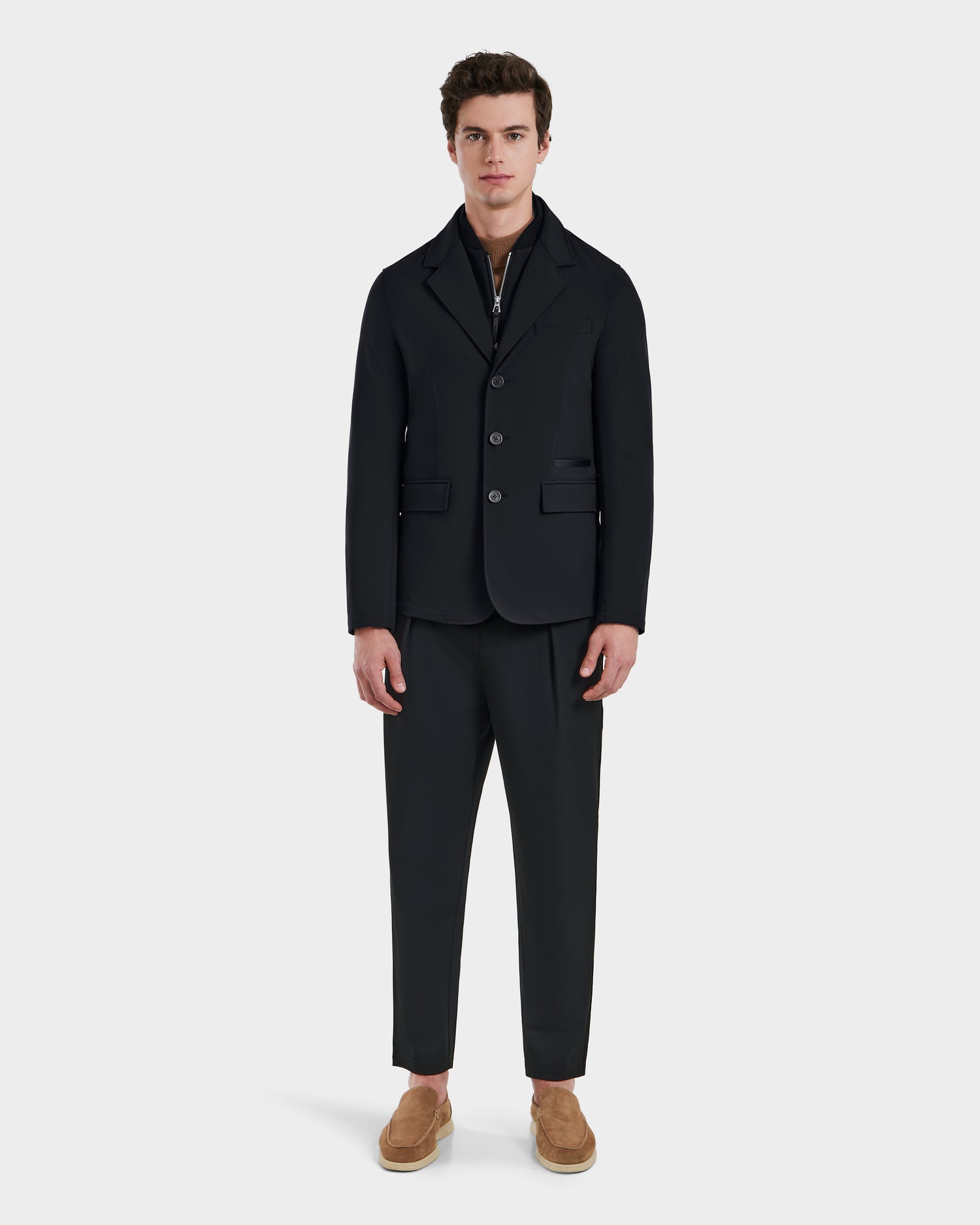 A man stands against a plain background, wearing Rudsak’s KEITH - 6125045 Black jacket with matching trousers, a zip-up shirt, and light brown slip-ons—a refined look that showcases modern tailoring in versatile menswear.