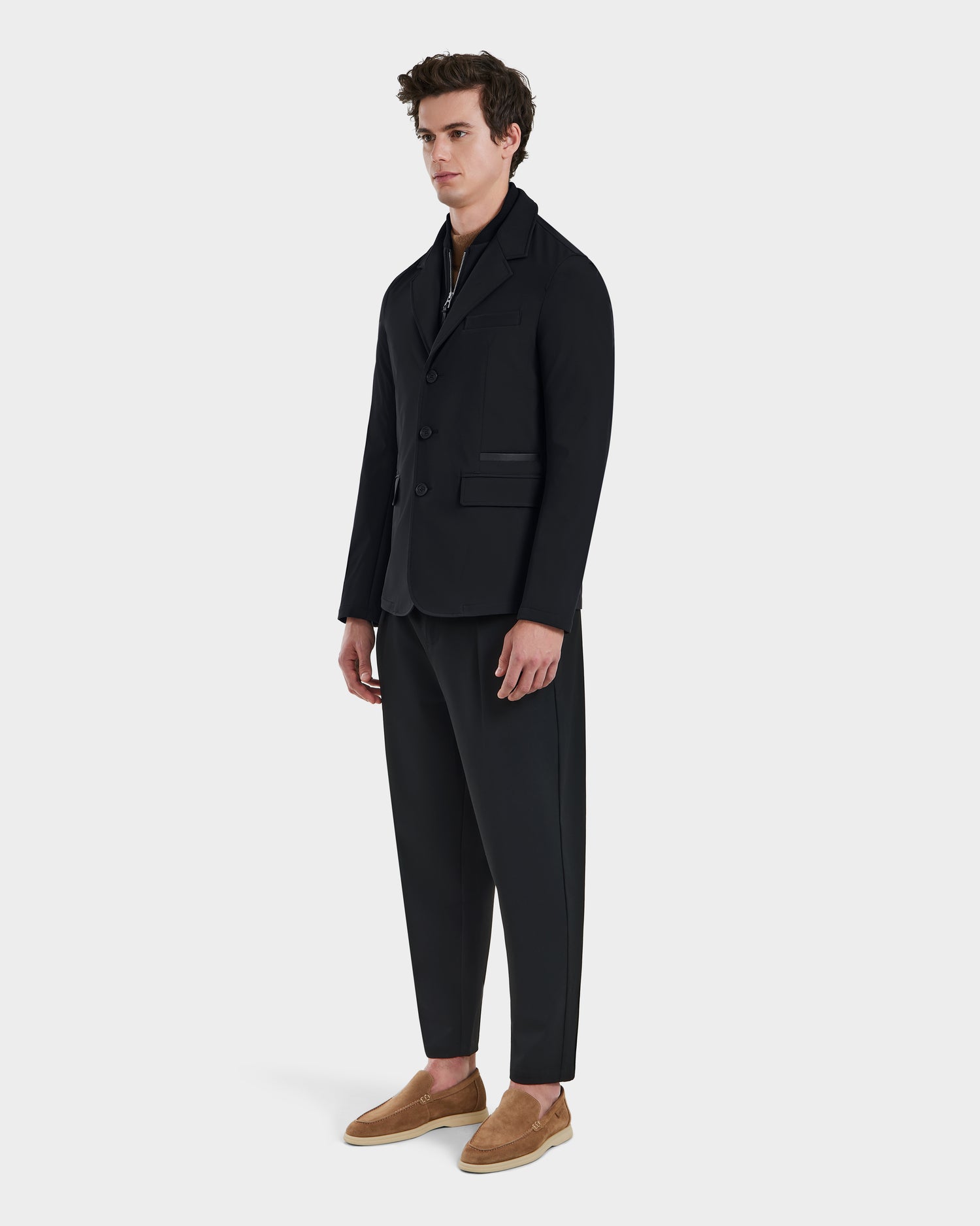 A man stands against a plain light background, wearing the Rudsak KEITH - 6125045 Black blazer-style jacket and matching trousers, paired with a dark shirt and tan slip-on shoes. He looks forward neutrally, showcasing modern tailoring.