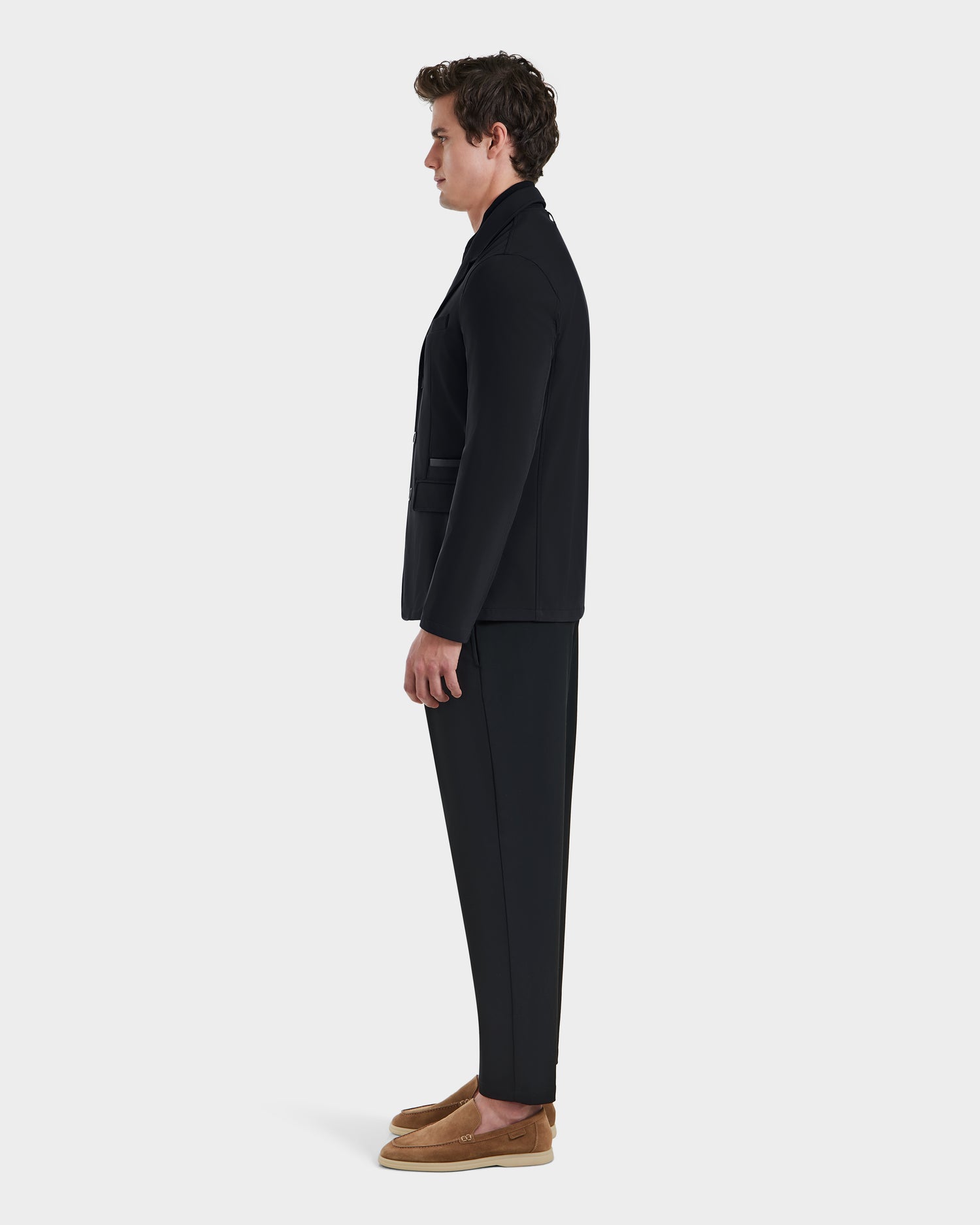 A man stands in profile facing left, wearing the Rudsak KEITH - 6125045 Black blazer-style jacket with a black suit and tan loafers, set against a plain white background.