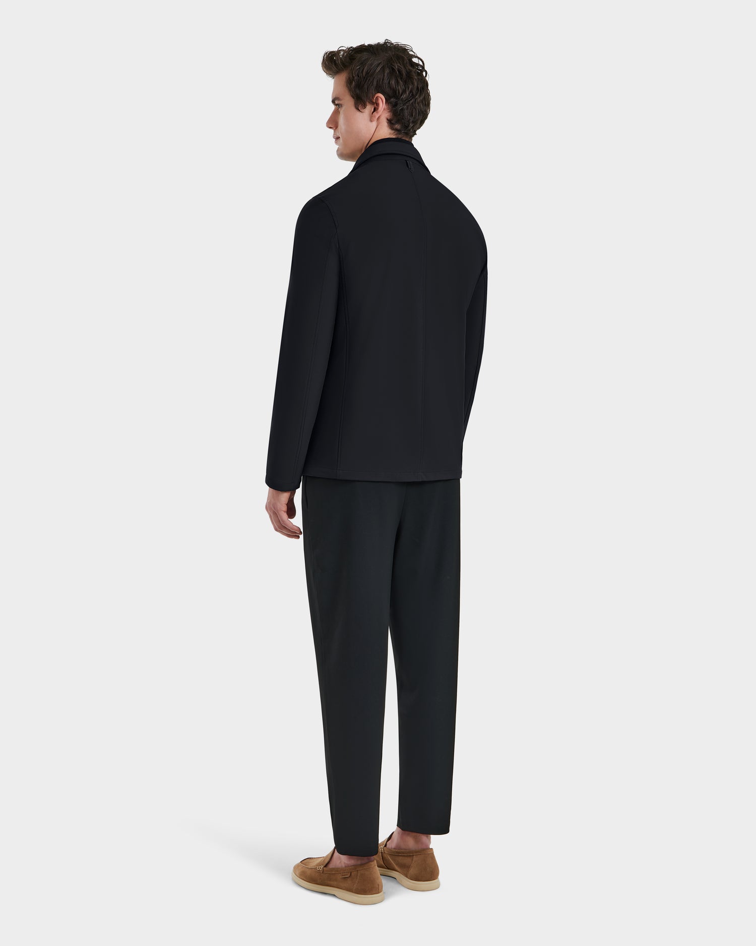 A man faces away wearing the Rudsak KEITH - 6125045 Black blazer-style jacket, paired with black pants and tan slip-on shoes, set against a plain light gray background—showcasing modern, versatile menswear.