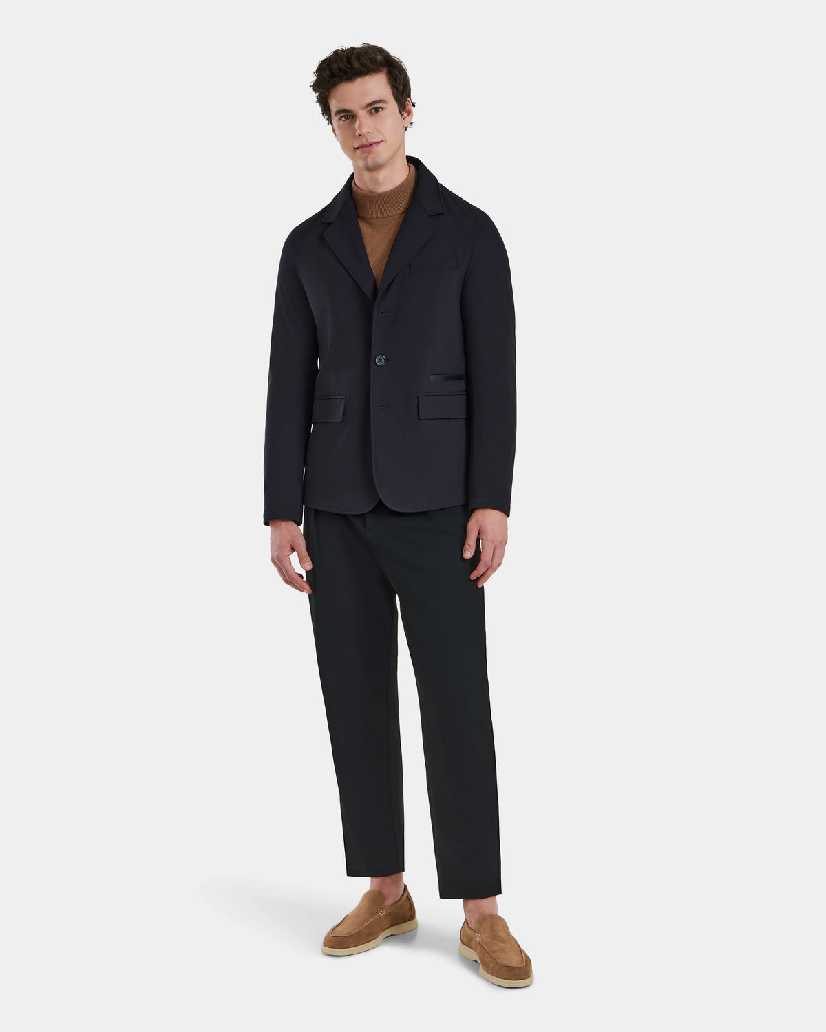 A man with short dark hair stands against a white background wearing the KEITH - 6125045 Black blazer-style jacket by Rudsak, paired with a brown turtleneck, black pants, and tan slip-on shoes for a modern, versatile menswear look.