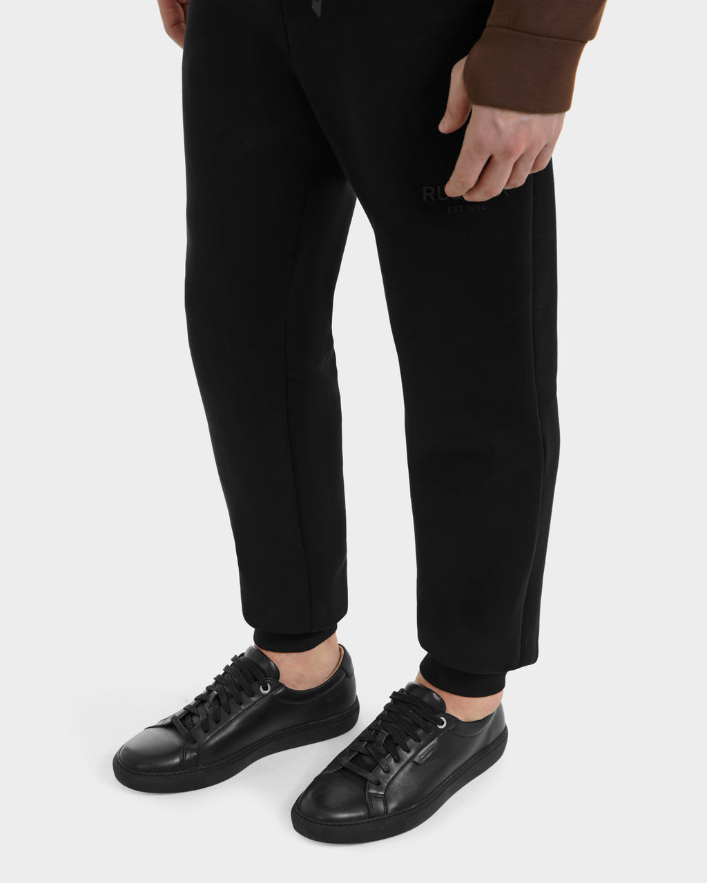A person in black LARIS - 6225000 jogger pants and Rudsak men’s leather sneakers stands on a light gray surface, with only their lower half and brown long-sleeve top visible.