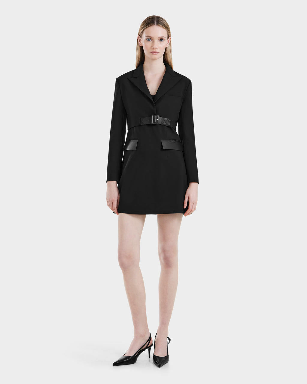 A woman stands against a white background wearing the Rudsak LEXI - 8525026 Black blazer dress with long sleeves, a belted waist, flap pockets, and black pointed-toe heels.