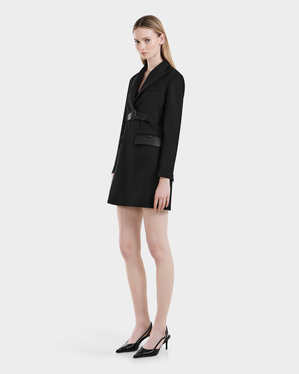 A woman wears the Rudsak LEXI - 8525026 Black vegan leather blazer dress with a belted waist and black pointed-toe heels, standing against a plain white background with a neutral expression.