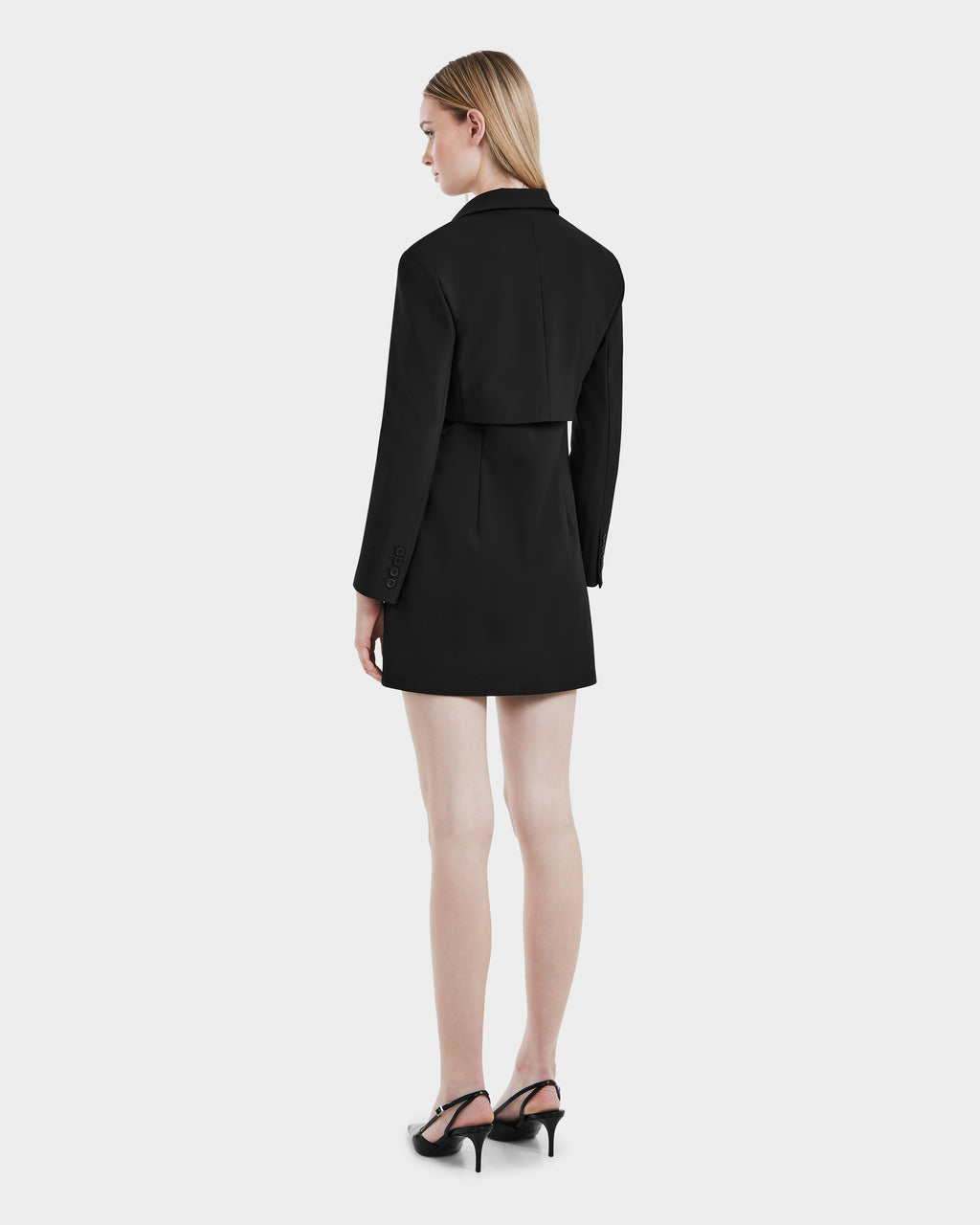 A woman with straight blonde hair stands facing away, wearing the Rudsak LEXI - 8525026 black vegan leather blazer dress with long sleeves and black heeled shoes against a plain light background.