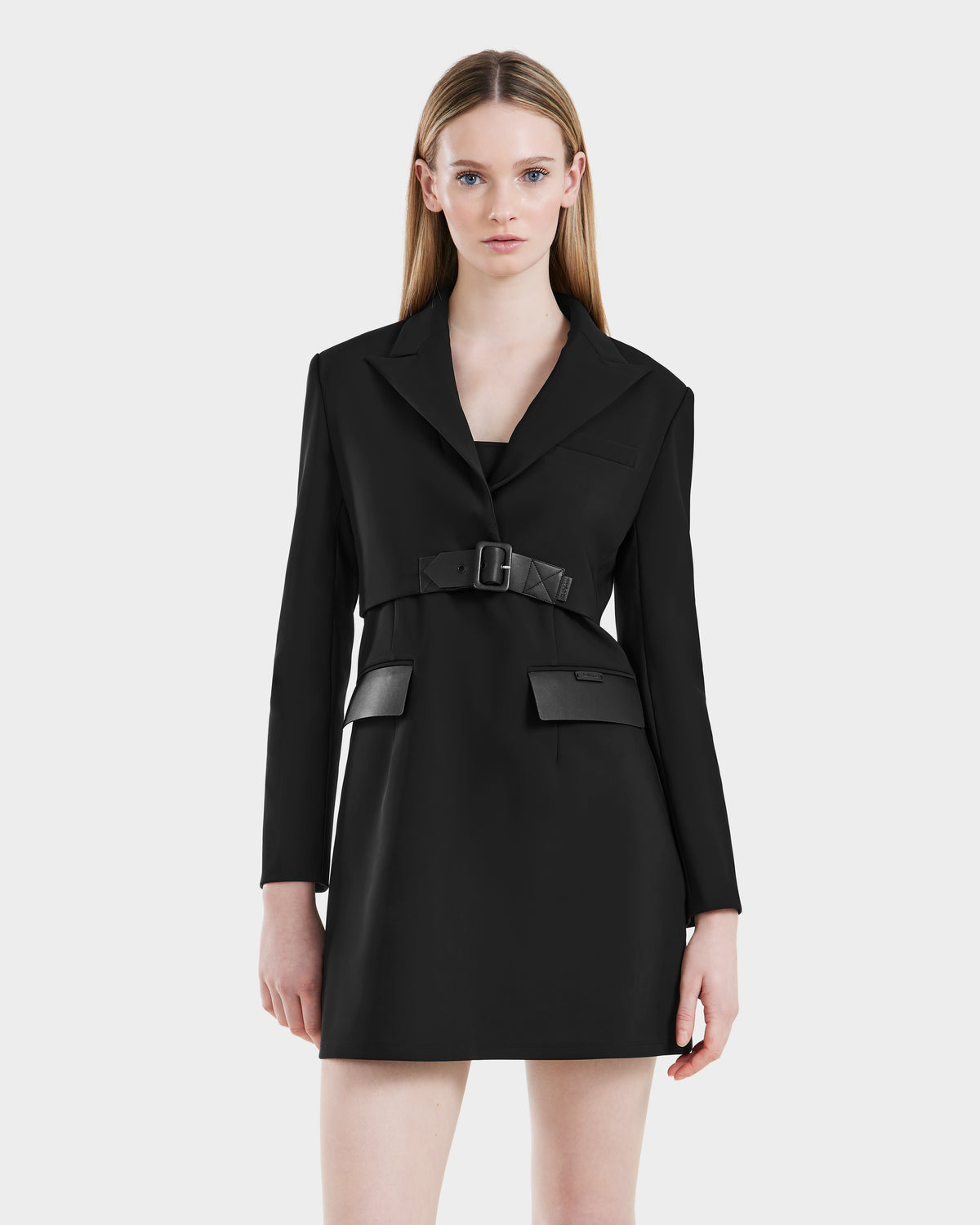 A woman with straight, light brown hair models the Rudsak LEXI - 8525026 Black tailored blazer dress with a vegan leather belt and flap pockets, standing against a white background—a modern take on power dressing.
