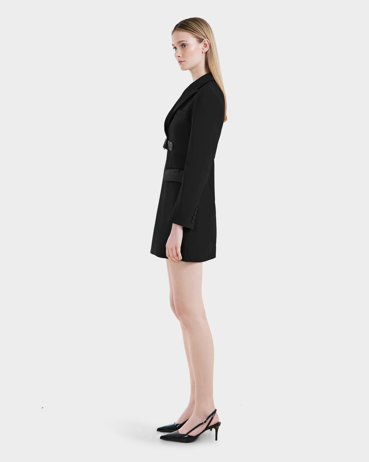 A woman with straight blonde hair stands in profile against a white background, wearing the Rudsak LEXI - 8525026 Black fitted blazer dress and black slingback heels.