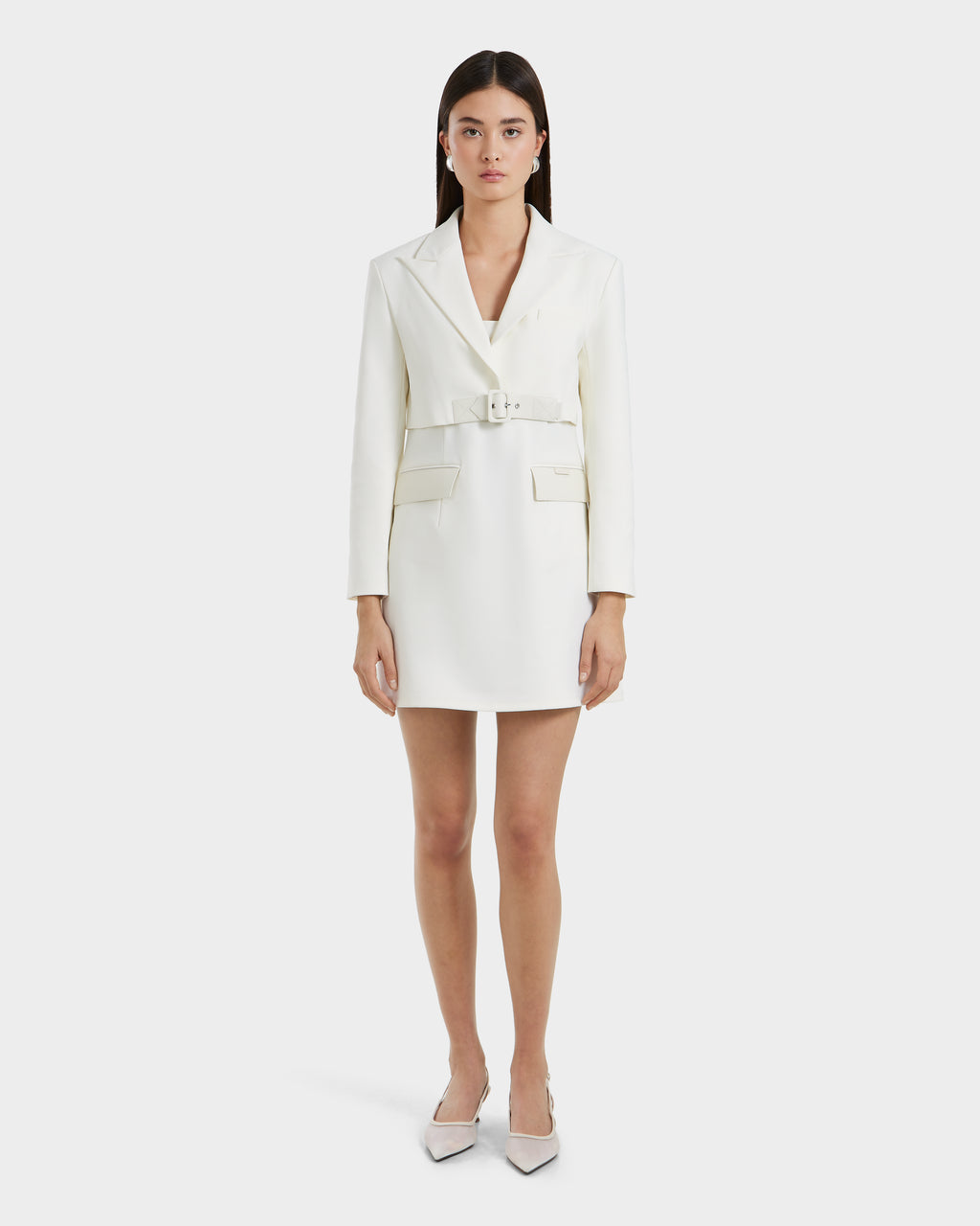 A woman showcases the Rudsak LEXI - 8525026 Iceland white blazer dress with a vegan leather belt, long sleeves, and front pockets, styled with pointed white heels for a sleek power look against a plain white backdrop.