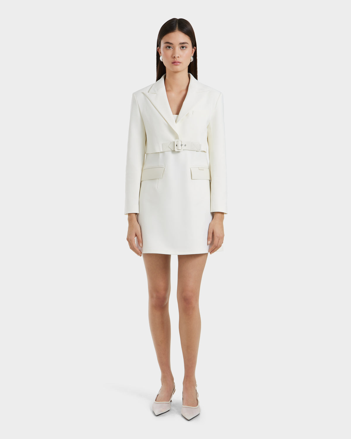 A woman showcases the Rudsak LEXI - 8525026 Iceland white blazer dress with a vegan leather belt, long sleeves, and front pockets, styled with pointed white heels for a sleek power look against a plain white backdrop.