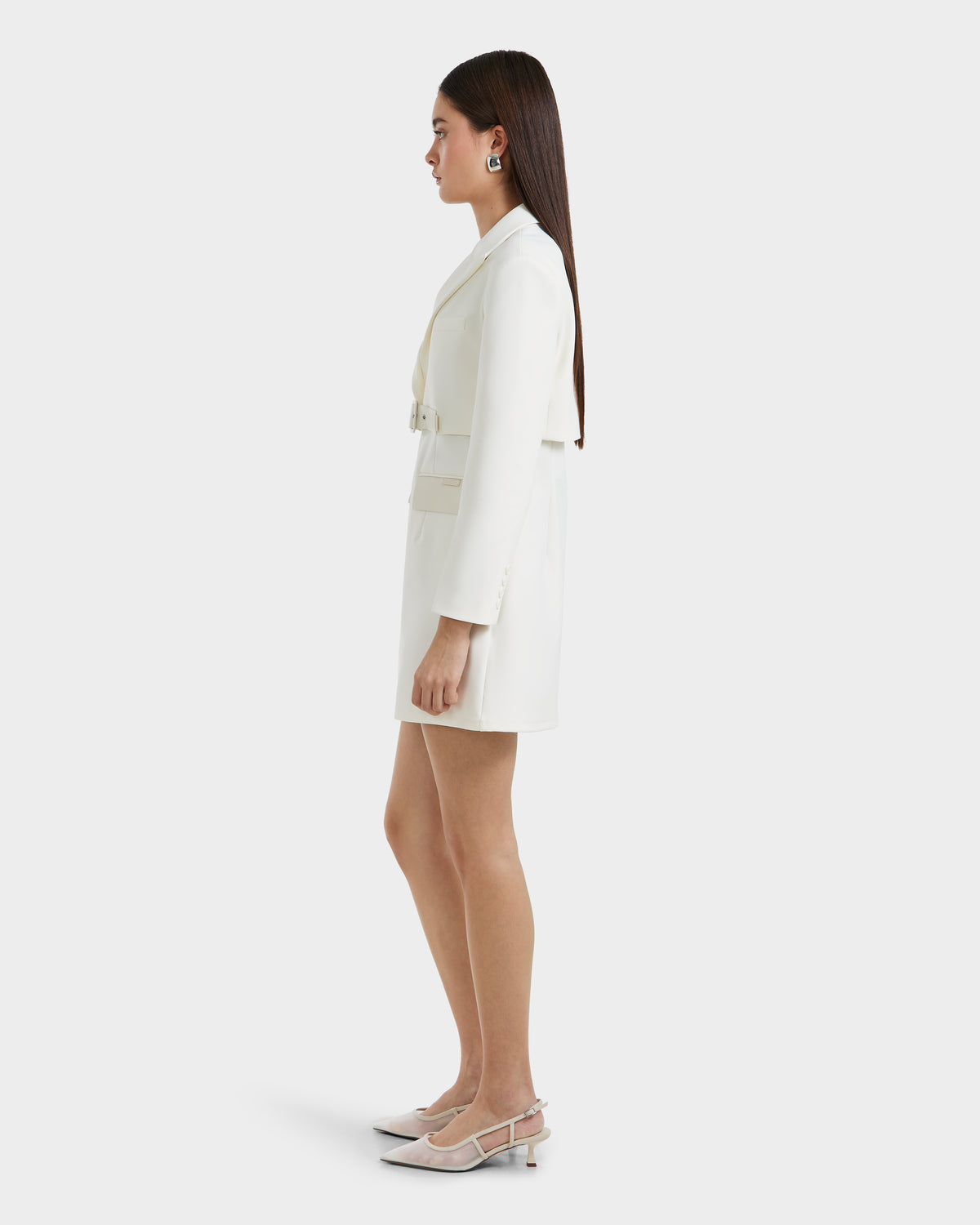 A woman with straight, long brown hair stands in profile, showcasing power dressing in Rudsaks LEXI - 8525026 Iceland white tailored blazer dress, cinched with a vegan leather belt and paired with beige heels against a plain white background.