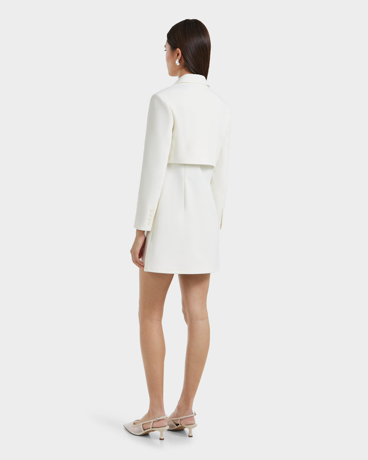 A woman with straight, dark hair stands against a light background, wearing the Rudsak LEXI - 8525026 Iceland white blazer dress with a vegan leather belt and beige heeled sandals. The dress features a cropped back overlay for a powerful look.