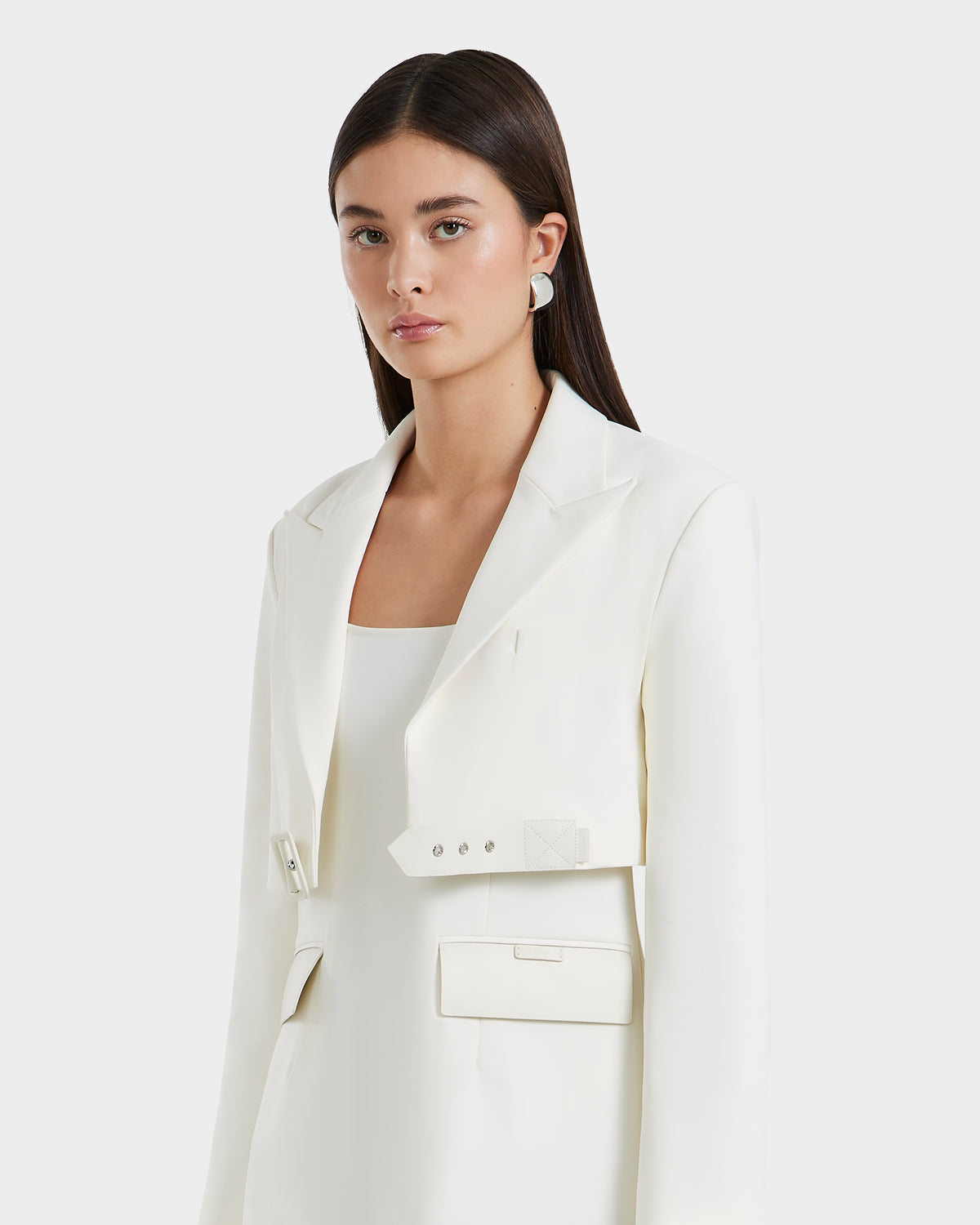 A woman with straight dark hair models the Rudsak LEXI - 8525026 Iceland cropped blazer, featuring distinctive cut-outs at the waist over a white top and vegan leather belt, standing against a plain light background.
