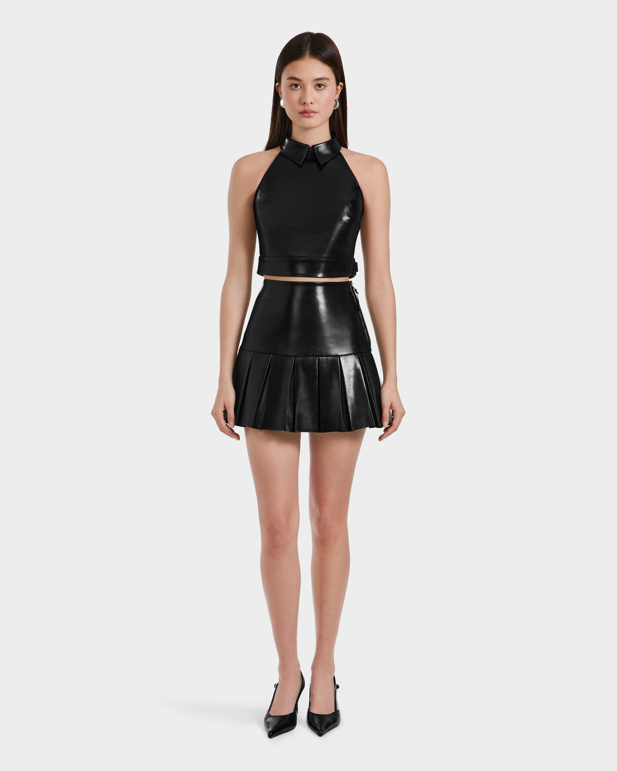 A woman with straight brown hair faces forward, wearing the Rudsak LOVELL - 8525557 Black leather sleeveless crop top with collar and matching high-waisted pleated mini skirt, paired with black pointed-toe heels against a plain white background.