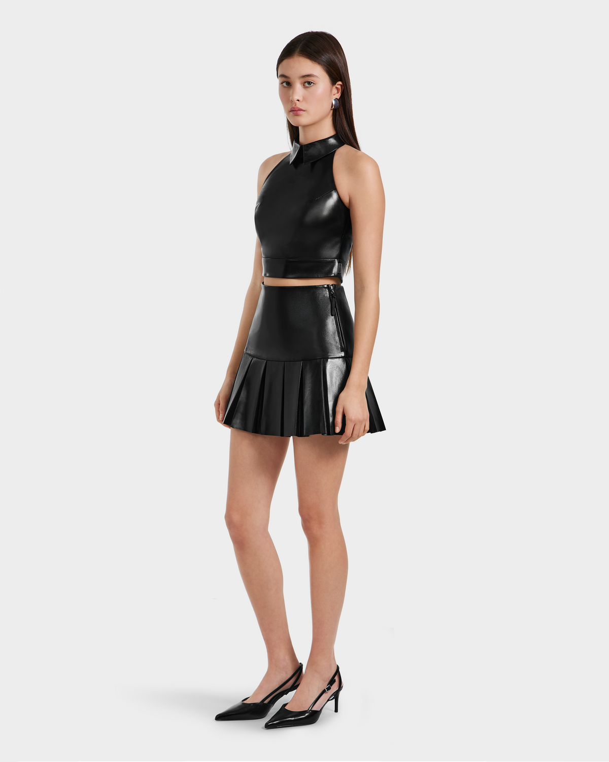 A woman wears the Rudsak LOVELL - 8525557 Black sleeveless leather top and matching pleated mini skirt with black pointed-toe heels against a plain light background.