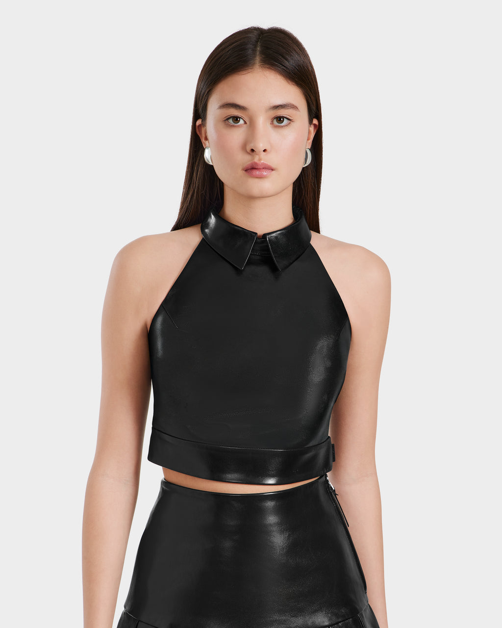 A woman with long brown hair models the Rudsak LOVELL - 8525557 Black leather halter top with collar and matching high-waisted skirt, standing against a plain light gray background.