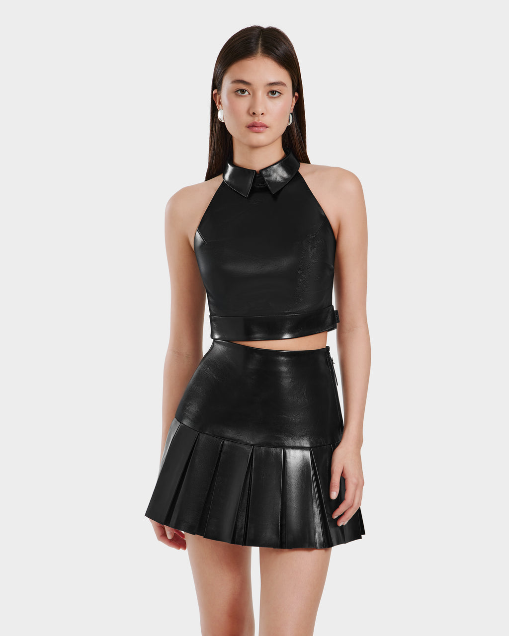 A young woman models the Rudsak LOVELL - 8525557 Black leather halter top with collar and matching high-waisted pleated mini skirt, standing against a plain white background.