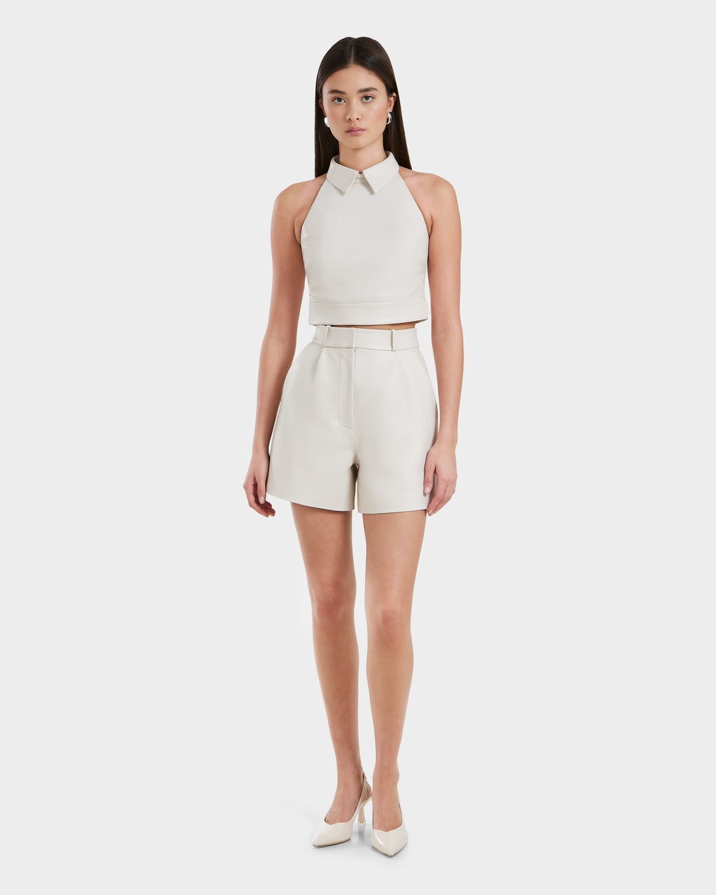 A woman faces forward wearing the Rudsak LOVELL - 8525557 Iceland women’s leather halter top in light beige, paired with high-waisted shorts and pointed-toe heels against a plain white background.