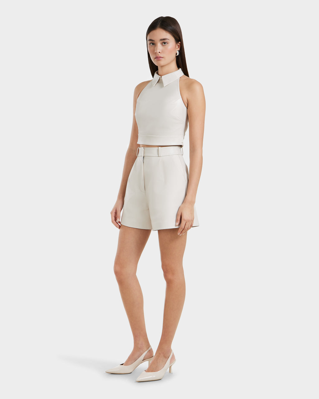 A woman wears the Rudsak LOVELL - 8525557 Iceland: an all-white premium leather sleeveless top with high-waisted belted shorts and white heels, posed against a plain white background.