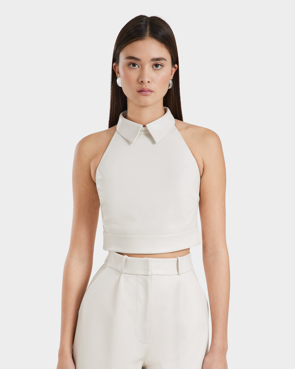 A woman with long dark hair models the Rudsak LOVELL - 8525557 Iceland sleeveless leather halter top with a collar, paired with high-waisted ivory wide-leg pants, standing against a plain white background.