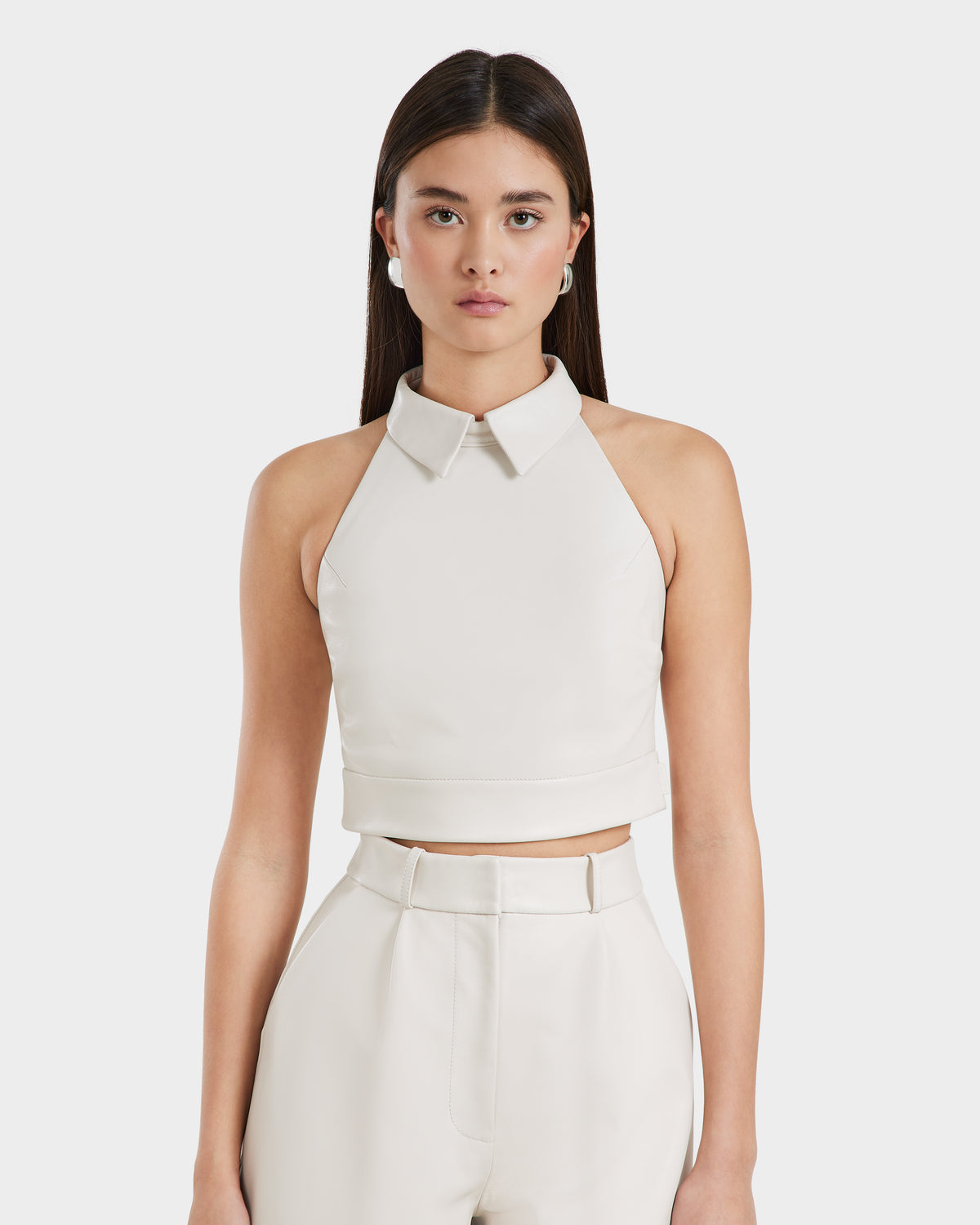 A woman with long dark hair models the Rudsak LOVELL - 8525557 Iceland sleeveless leather halter top with a collar, paired with high-waisted ivory wide-leg pants, standing against a plain white background.